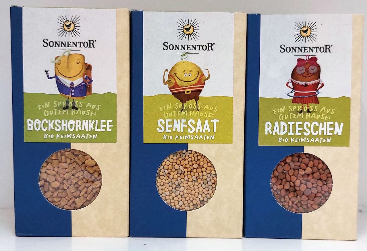 Drei Schachteln mit Bio-Saatgut sind ausgestellt. Die linke Schachtel ist mit 'SHORNKLEE' beschriftet, die mittlere mit 'SENFSAAT' und die rechte mit 'RADIES'. Jede Schachtel zeigt eine Cartoon-Figur und einen Kreis aus Samen.