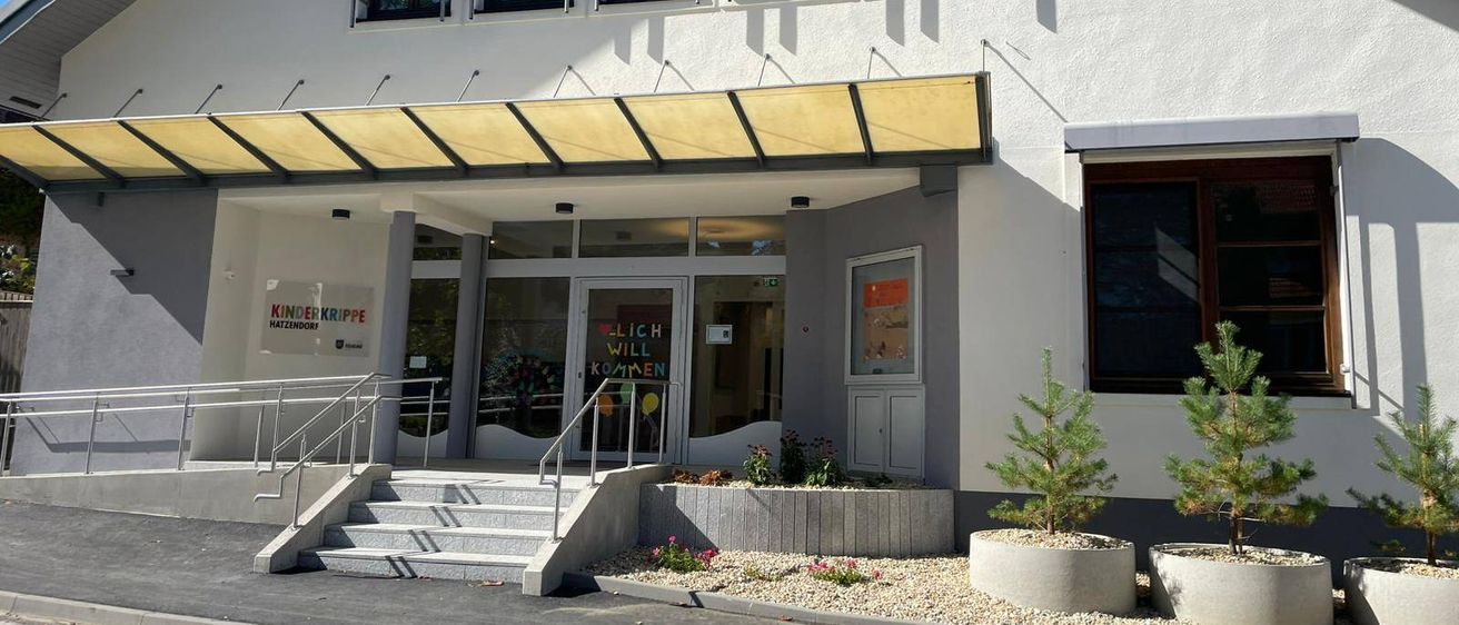 Ein weißes Gebäude mit drei Fenstern und einer Tür. Der Eingang hat ein Schild, das 'Kinderkrippe Nattendorf' besagt. Es gibt eine Treppe mit Geländern, die zum Eingang führt. Zwei Topfpflanzen befinden sich an der Seite.