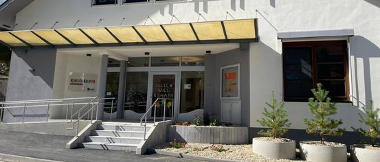 Ein weißes Gebäude mit drei Fenstern und einer Tür. Der Eingang hat ein Schild, das 'Kinderkrippe Nattendorf' besagt. Es gibt eine Treppe mit Geländern, die zum Eingang führt. Zwei Topfpflanzen befinden sich an der Seite.