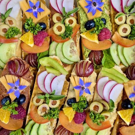 Eine Auswahl an Sandwiches mit verschiedenen Belägen wie Käse, Früchten, Gemüse und Blumen, die auf ansprechende Weise angeordnet sind.