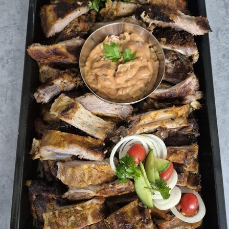 Ein Tablett mit geräucherten Rippchen, einer Schüssel cremiger Sauce, garniert mit Petersilie und begleitet von geschnittenen Zwiebeln und Tomaten.
