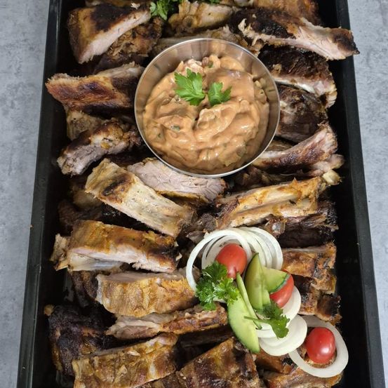 Ein Tablett mit geräucherten Rippchen, einer Schüssel cremiger Sauce, garniert mit Petersilie und begleitet von geschnittenen Zwiebeln und Tomaten.