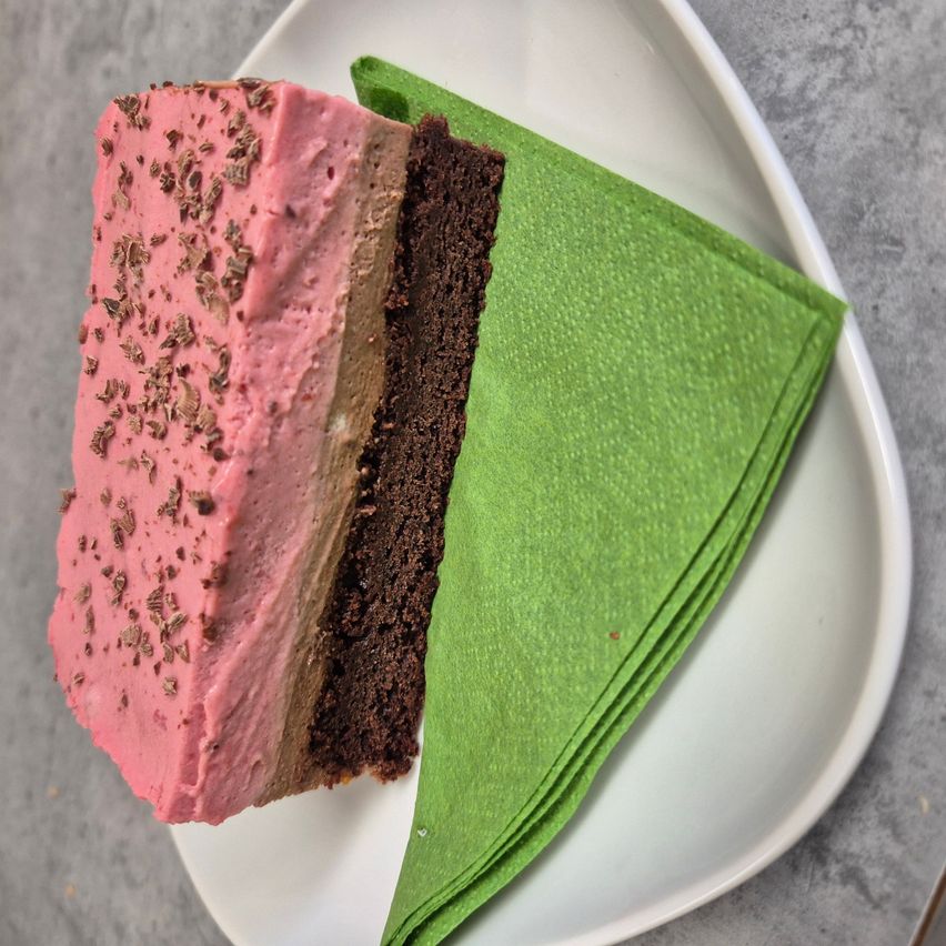 Eine Scheibe mehrschichtiger Kuchen mit rosa Glasur und Schokoladenraspeln auf einem weißen Teller, begleitet von grünen Servietten.