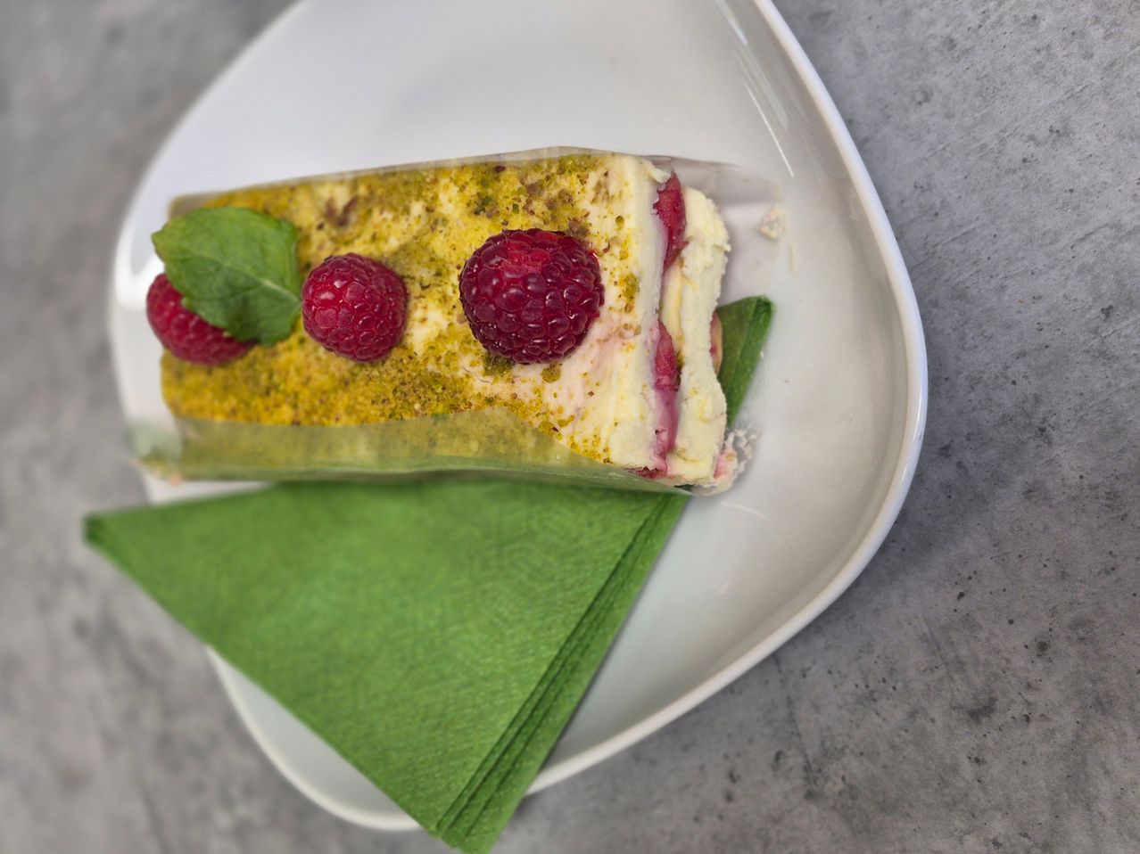 Ein Stück Pistazien-Himbeer-Kuchen auf einem weißen Teller, garniert mit drei Himbeeren und einem Minzblatt.