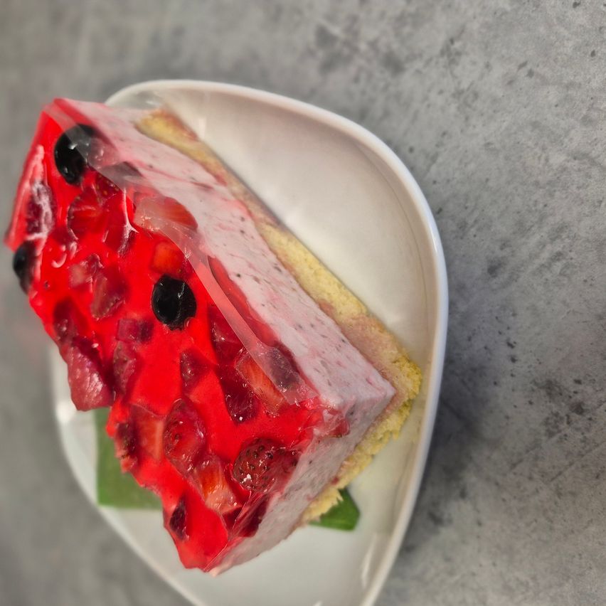 Ein Stück Erdbeer-Gelee-Kuchen auf einem weißen Teller, mit grünen Blättern garniert, auf einer grauen Oberfläche.