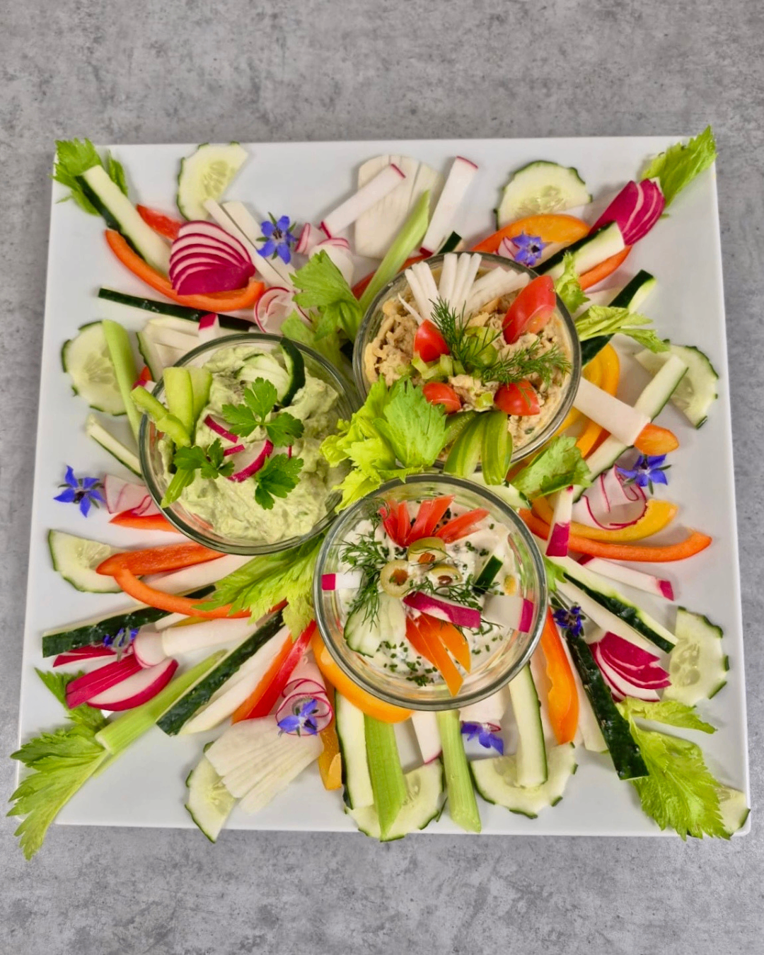 Ein weißer Teller mit einer Vielzahl von bunten Gemüsesorten und drei kleinen Dipschüsseln. Das Gemüse umfasst geschnittene Gurken, Karotten, Radieschen und Salat. Die Dips sind grün, weiß und rot.