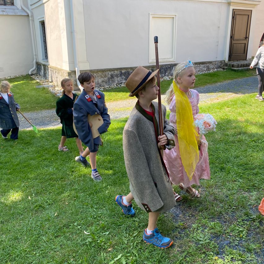 Eine Gruppe von Kindern in Kostümen geht auf einem Rasen. Einer hält einen Stab, ein anderer eine Blumenstrauß. Dahinter steht ein Gebäude mit einer großen Tür und Fenstern.