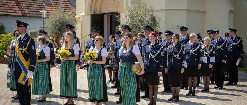 Eine Gruppe von Menschen in Uniformen und traditionellen Kleidern steht in einer Reihe vor einem Gebäude. Einige halten gelbe Blumen und Musikinstrumente.