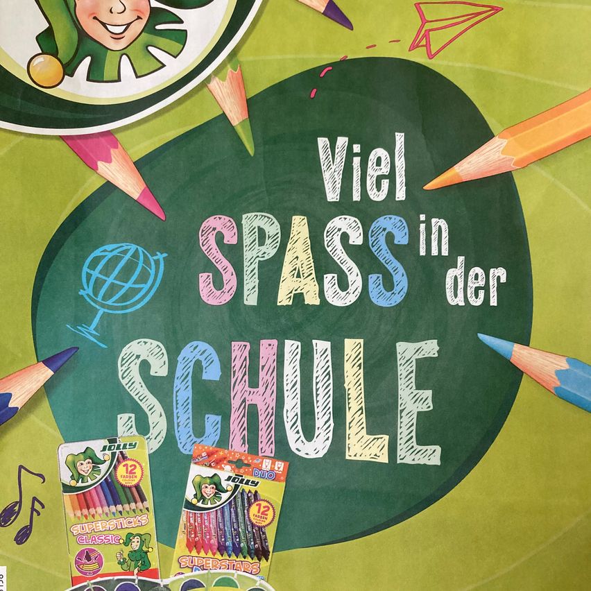 Eine Tafel mit 'Viel Spass in der Schule' in bunter Kreide geschrieben. Drumherum sind Stifte, Papierflugzeuge und ein Globus. Unten an der Tafel liegen zwei Packungen Buntstifte, eine beschriftet mit 'Jolly Supersticks Classic' und die andere mit 'Jolly Duo Superstars'.