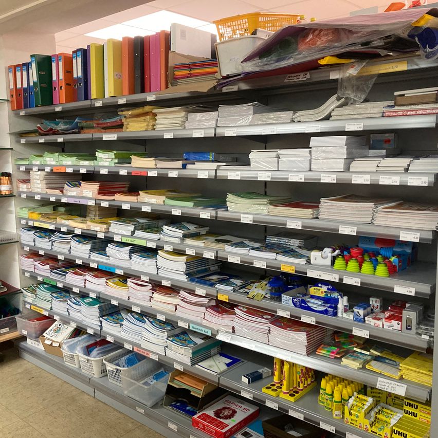 Ein Ladenregal voller verschiedener Büroartikel. Die Regale sind mit Ordnern, Heftern, Notizbüchern und verschiedenen anderen Schreibwaren bestückt. Es gibt auch Aufbewahrungsboxen und Behälter. Die Decke hat ein helles Licht, das den Bereich beleuchtet.