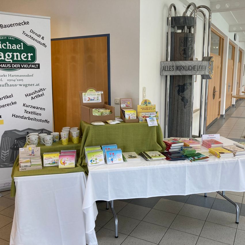 Zwei Tische mit weißen Tischdecken sind in einem Raum aufgestellt und zeigen eine Vielzahl von Artikeln. Ein Tisch hat Bücher, Notizbücher und Tassen, während der andere eine Schachtel mit grünen Artikeln hat. Hinter den Tischen befindet sich eine Holztür, und ein Schild steht 'ALLES WIRKLICHE LEBEN.' Ein Werbebanner ist links.