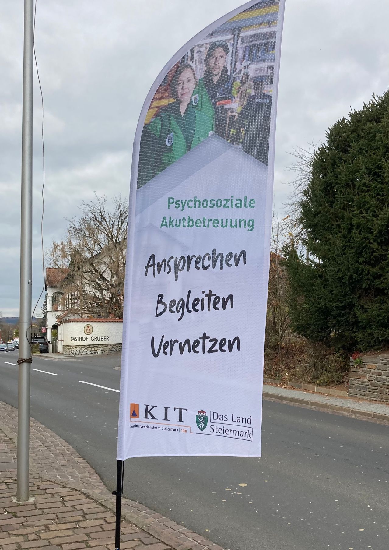 Ein weißer Banner mit einem Foto von Personen in Uniformen und dem Text 'Psychosoziale Akutbetreuung Ansprechen Begleiten Vernetzen' hängt an einer Straße. Unten sind Logos für KIT und Das Land Steiermark zu sehen. Im Hintergrund befindet sich eine Straße mit Autos und ein Gebäude.