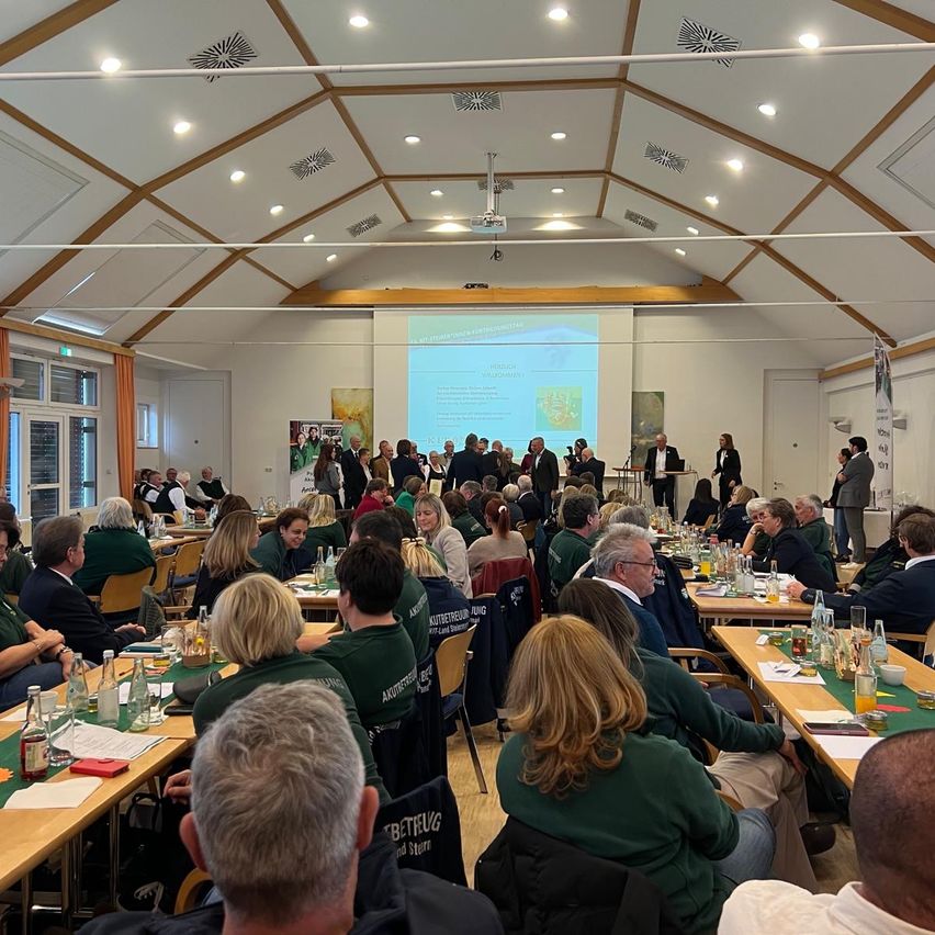 Ein Konferenzsaal mit einer Projektionsleinwand, Menschen an Tischen mit Flaschen und Papieren, und einem Redner vorne. Einige Personen stehen in der Nähe des Eingangs.