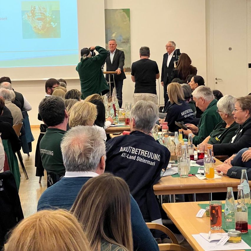 Eine Gruppe von Menschen versammelt sich in einem Konferenzraum. Ein Mann steht an einem Podium mit einer Projektionsleinwand dahinter. Mehrere Teilnehmer sitzen an Tischen mit Getränken und Snacks. Eine Frau in einem grünen Shirt hält das Ereignis mit einer Kamera fest.