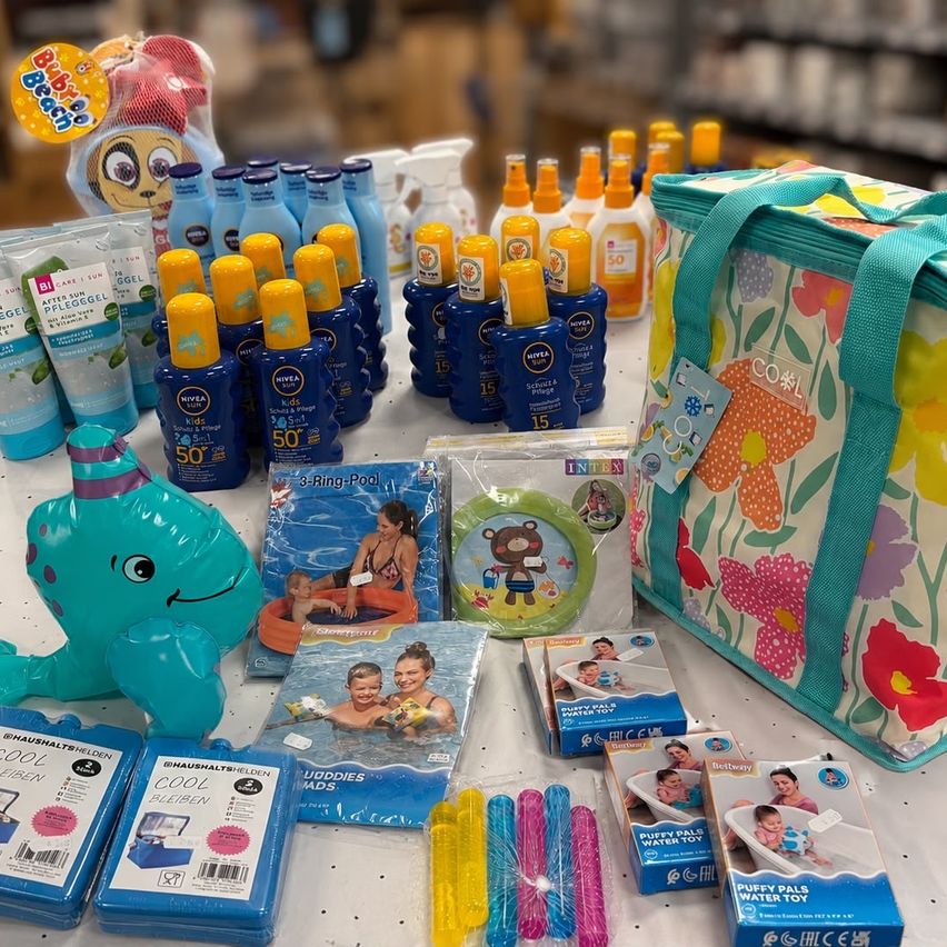 Ein Tisch zeigt verschiedene Poolzubehör für Kinder, darunter aufblasbare Spielzeuge, Sonnencremeflaschen und eine Blumen-Lunchtasche. Die Artikel sind ordentlich angeordnet, mit einem Cartoon-Zeichentrickfiguren-Spielzeug und einem kleinen Pool im Hintergrund.