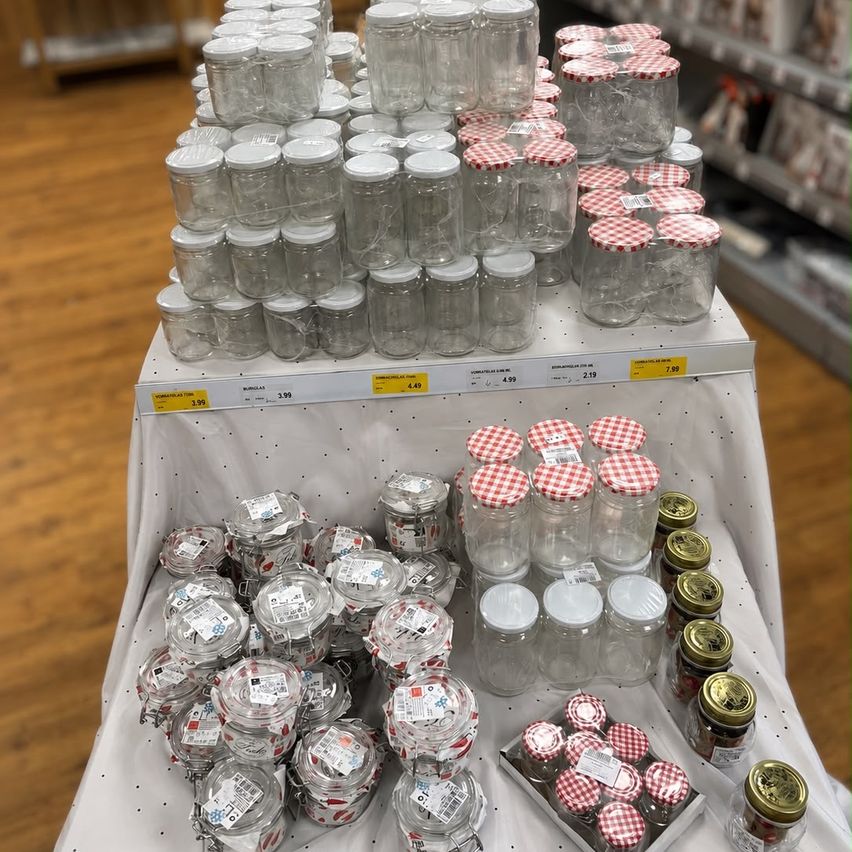 A store display of various glass jars with red and white checkered lids, some stacked and others on a white cloth-covered table, with price tags visible.