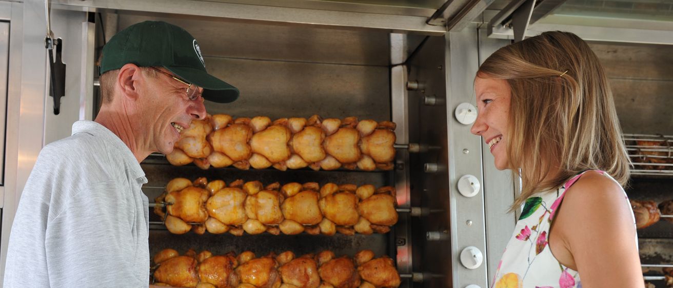 Ein Mann und eine Frau lächeln und schauen auf einen Grill voller Hühnchen. Der Mann hält einen Stock mit Hühnchen daran.
