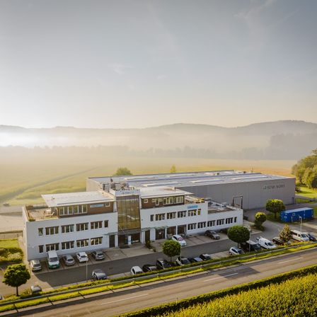 Luftaufnahme eines großen Industriekomplexes mit mehreren Gebäuden, umgeben von einem Parkplatz und Grünflächen, vor dem Hintergrund von nebeligen Bergen.