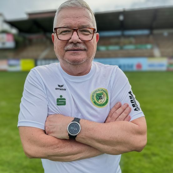 Ein älterer Mann steht mit verschränkten Armen auf einem Rasenfeld, trägt ein weißes Shirt mit Logo und eine Uhr. Im Hintergrund ist ein leeres Stadion zu sehen.