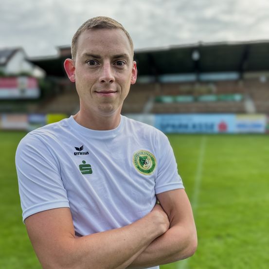 Ein lächelnder Mann steht mit verschränkten Armen auf einem Fußballfeld. Dahinter ist ein verschwommener Blick auf ein leeres Stadion zu sehen.