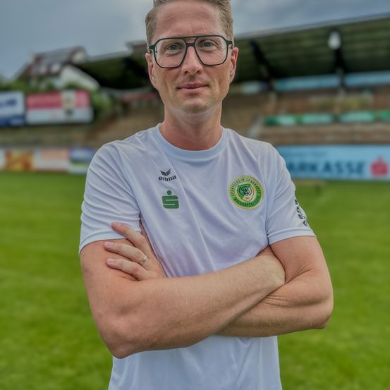 Ein Mann in einem weißen T-Shirt steht auf einem grünen Feld mit verschränkten Armen und trägt eine Brille. Hinter ihm ist eine verschwommene Ansicht eines Stadions mit leeren Tribünen.