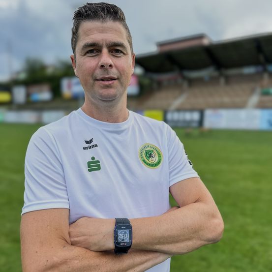 Ein Mann steht mit verschränkten Armen auf einem Fußballfeld. Er trägt ein weißes Shirt mit Logo und Uhr. Im Hintergrund ist ein leeres Stadion mit einem grünen Feld zu sehen.