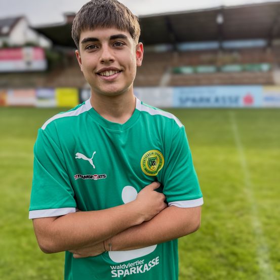 Ein junger Mann steht auf einem Fußballplatz und lächelt mit verschränkten Armen. Er trägt ein grünes Trikot mit einem weißen Logo auf der Brust. Im Hintergrund ist ein Stadion mit leeren Tribünen und einem Sponsorenschild zu sehen.