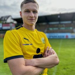 Ein Mann in einem gelben Fußballtrikot steht mit verschränkten Armen lächelnd auf einem Feld. Hinter ihm befindet sich ein Stadion mit einem sichtbaren Haus.