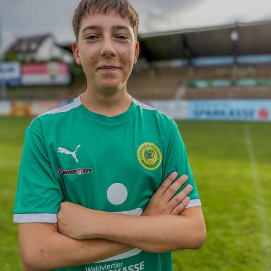 Ein junger Mann in einem grünen Sporttrikot mit Logo steht auf einem Fußballfeld, Arme verschränkt, mit einem Stadion und Häusern im Hintergrund.