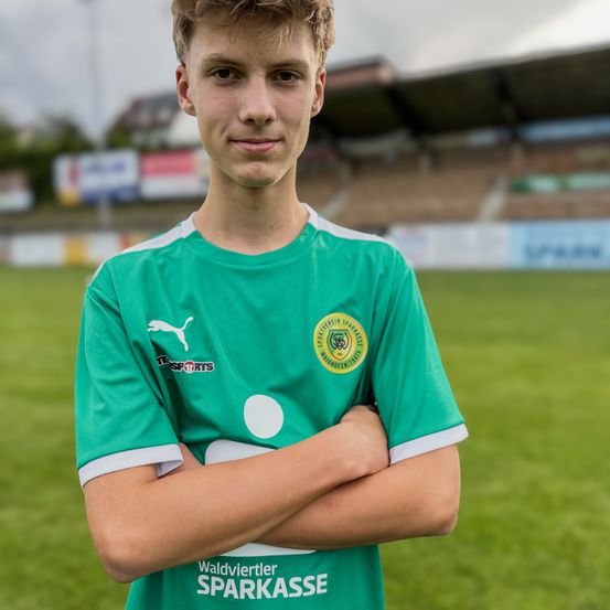 Ein junger männlicher Fußballspieler steht vor einem grünen Feld. Er trägt ein grünes Trikot mit weißen Akzenten. Er lächelt und verschränkt die Arme vor sich.