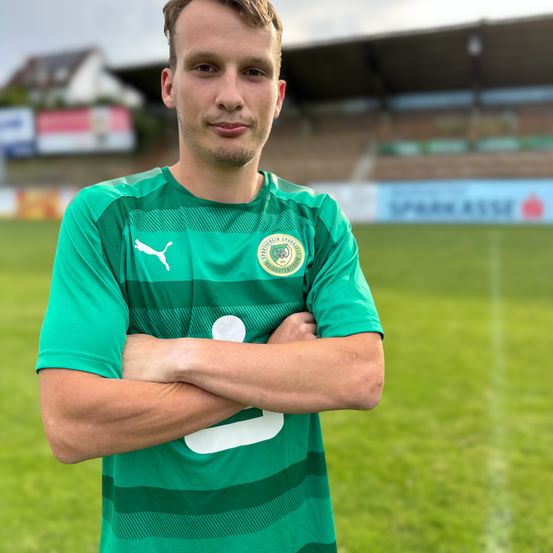 Ein Fußballspieler in einem grünen Trikot mit Logo steht auf einem Feld mit verschränkten Armen. Im Hintergrund sieht man ein Stadion mit leeren Tribünen und einem verschwommenen Banner.