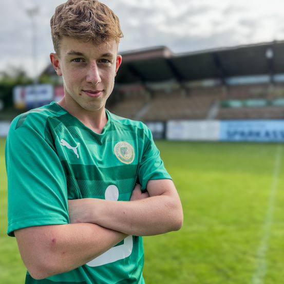 Ein junger männlicher Fußballspieler steht mit gefalteten Armen auf einem Feld und trägt ein grünes Trikot.