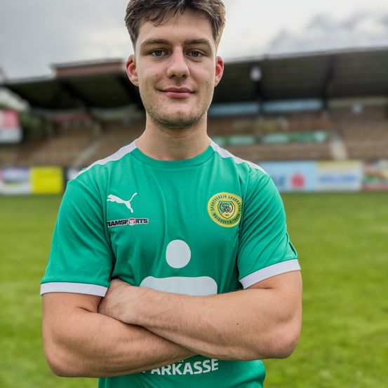Ein lächelnder Mann steht auf einem Fußballfeld mit verschränkten Armen und trägt ein grünes Trikot mit weißen Kanten und einem gelben Emblem. Hinter ihm ist ein leeres Stadion zu sehen, und der Himmel ist teilweise bewölkt.