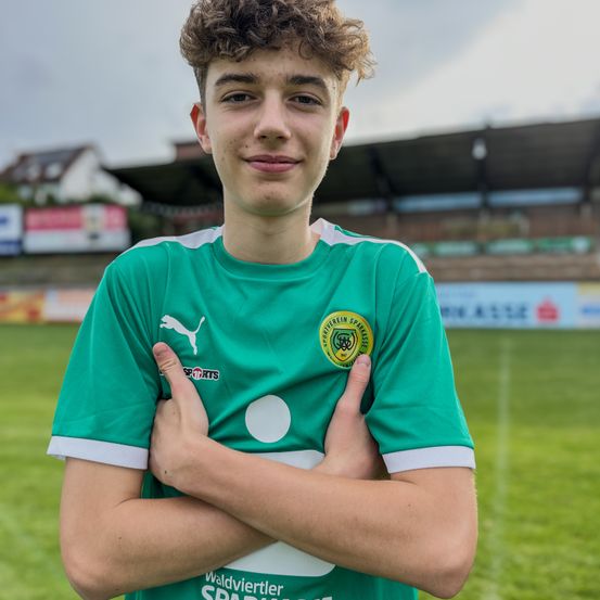 Ein junger Mann in einem grünen Fußballtrikot posiert mit verschränkten Armen vor einem Fußballfeld.