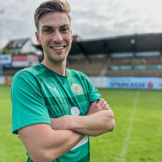 Ein Fußballspieler mit verschränkten Armen, lächelnd, steht vor einem Stadion. Er trägt ein grünes Trikot mit Logo auf der Brust.
