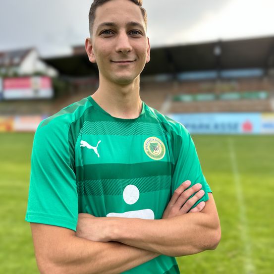 Ein Fußballspieler in einem grünen Trikot mit einem weißen Logo posiert für ein Foto auf einem Fußballfeld mit einem leeren Stadion im Hintergrund.