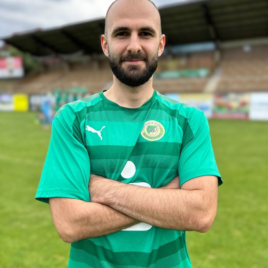 Ein Mann in einem grünen Fußballtrikot steht mit verschränkten Armen auf einem Feld und lächelt in die Kamera. Im Hintergrund ist eine verschwommene Ansicht eines leeren Stadions zu sehen.