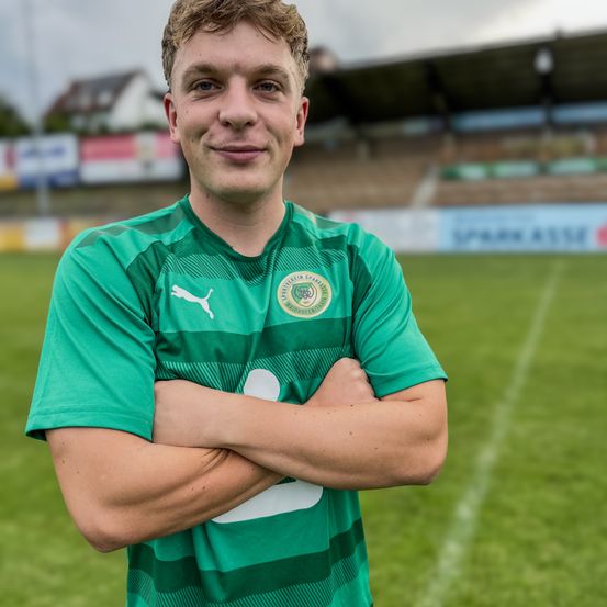 Ein Fußballspieler in einem grünen Trikot steht lächelnd auf dem Feld, die Arme verschränkt. Das Feld ist von leeren Tribünen umgeben, und ein blauer Banner ist im Hintergrund sichtbar.