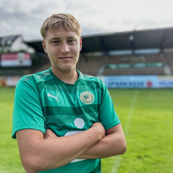 Ein junger Mann steht auf einem Fußballfeld und trägt ein grünes Trikot mit einem Logo auf der Brust. Er posiert mit verschränkten Armen, und hinter ihm ist ein verschwommenes Stadion mit Werbung.