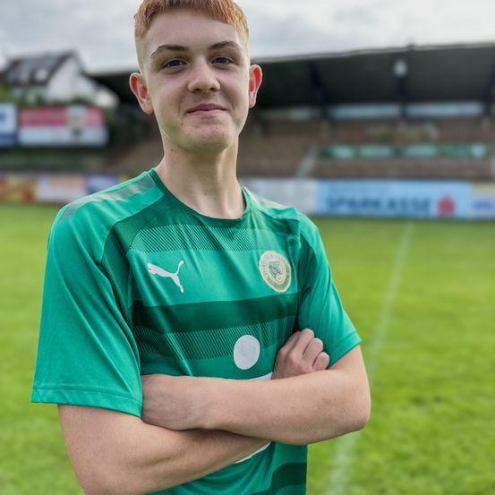 Ein junger Mann in einem grünen Fußballtrikot steht lächelnd auf einem Feld mit gefalteten Armen. Das Feld hat weiße Markierungen, und in der Ferne befindet sich ein verschwommenes Stadion mit Werbetafeln.