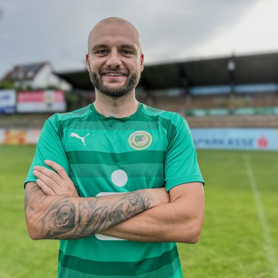 Ein kahlköpfiger Mann in einem grünen Fußballtrikot mit Logo steht lächelnd auf einem Feld mit verschränkten Armen. Dahinter befindet sich ein verschwommenes Stadion mit einem Rasenfeld.