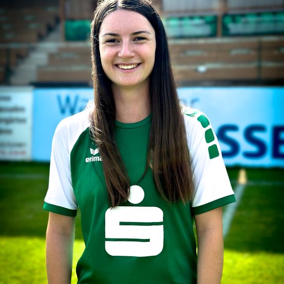 Eine Frau in einem grünen und weißen Fußballtrikot lächelt und steht auf einem Feld. Der Hintergrund zeigt ein Stadion mit Tribünen und einem Banner.