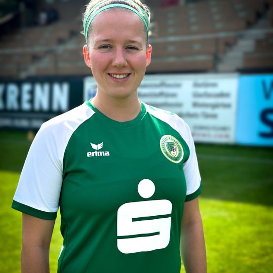 Eine Frau steht auf einem Rasenfeld und trägt ein grünes und weißes Fußballtrikot mit der Nummer '9'. Sie lächelt. Im Hintergrund ist ein verschwommener Hintergrund mit Text und Logo.