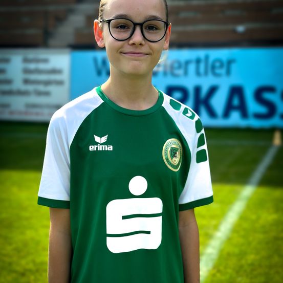 Ein junger Fußballspieler in einem grün-weißen Trikot mit dem Wort 'erima' und einem Wappen auf der Brust lächelt auf einem Fußballfeld. Hinter ihm befindet sich ein Banner mit den Worten 'Werlte' und 'ERKAS'.