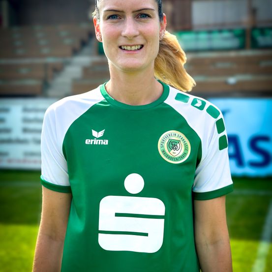 Eine Frau in einem grün-weißen Fußballtrikot lächelt für die Kamera auf einem Sportplatz mit verwaschenem Hintergrund.