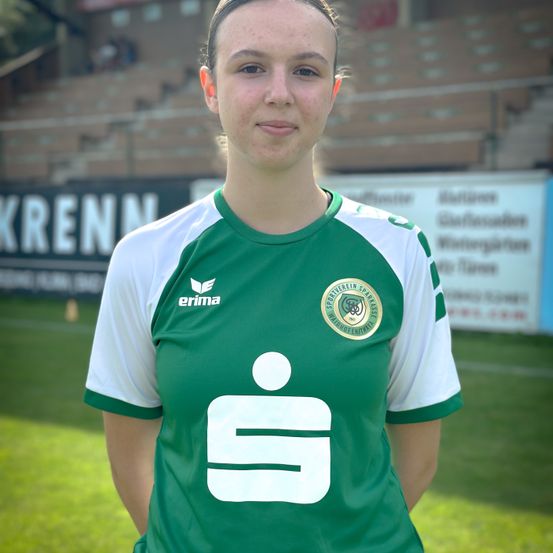 Eine Frau steht auf einem Fußballfeld, trägt ein grün-weißes Fußballtrikot mit der Nummer 18 und lächelt für ein Foto.