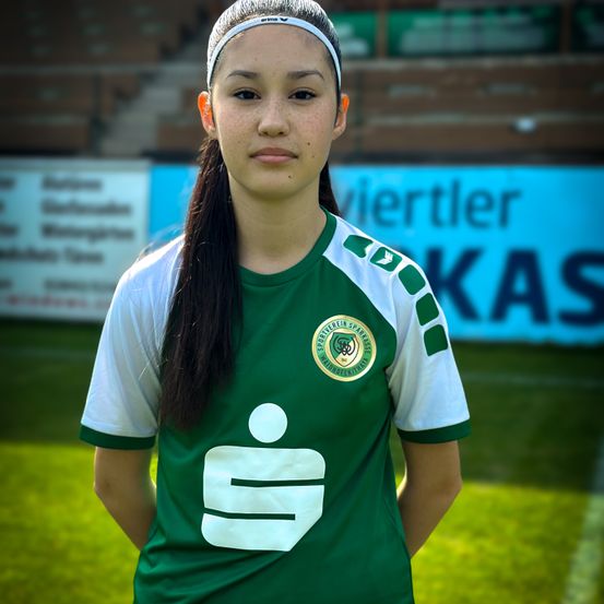 Eine junge Frau in einem grünen und weißen Sporttrikot mit Logo steht auf einem Fußballfeld mit unscharfem Hintergrund.