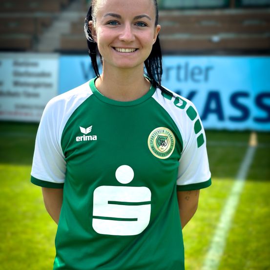 Eine lächelnde Frau in einem grünen und weißen Fußballtrikot mit der Nummer 9 steht auf einem Feld.