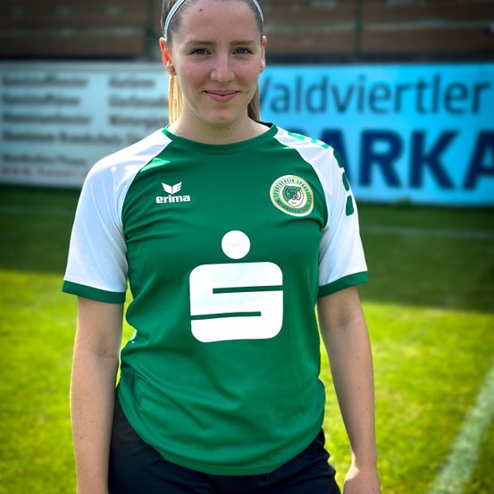 Eine Frau steht auf einem grünen Feld und trägt ein grünes Fußballtrikot mit einem Logo und dem Wort 'erima' darauf. Sie lächelt. Hinter ihr ist ein verschwommener Hintergrund mit einer Wand und einem Schild mit dem Wort 'ARKA'.