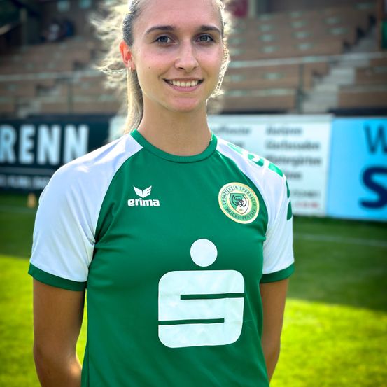 Eine lächelnde Frau in einem grünen und weißen Fußballtrikot mit einem Logo auf der Brust steht auf einem grünen Feld. Der Hintergrund zeigt leere Tribünen und Werbetafeln.